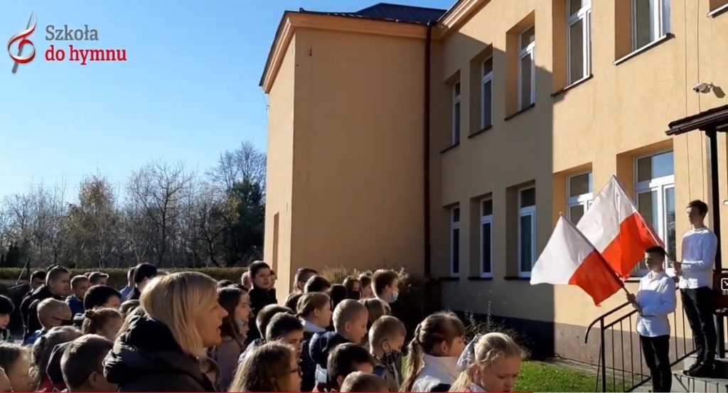 Uczniowie i nauczyciele spiewaja hymn przed budynkiem szkoły.
