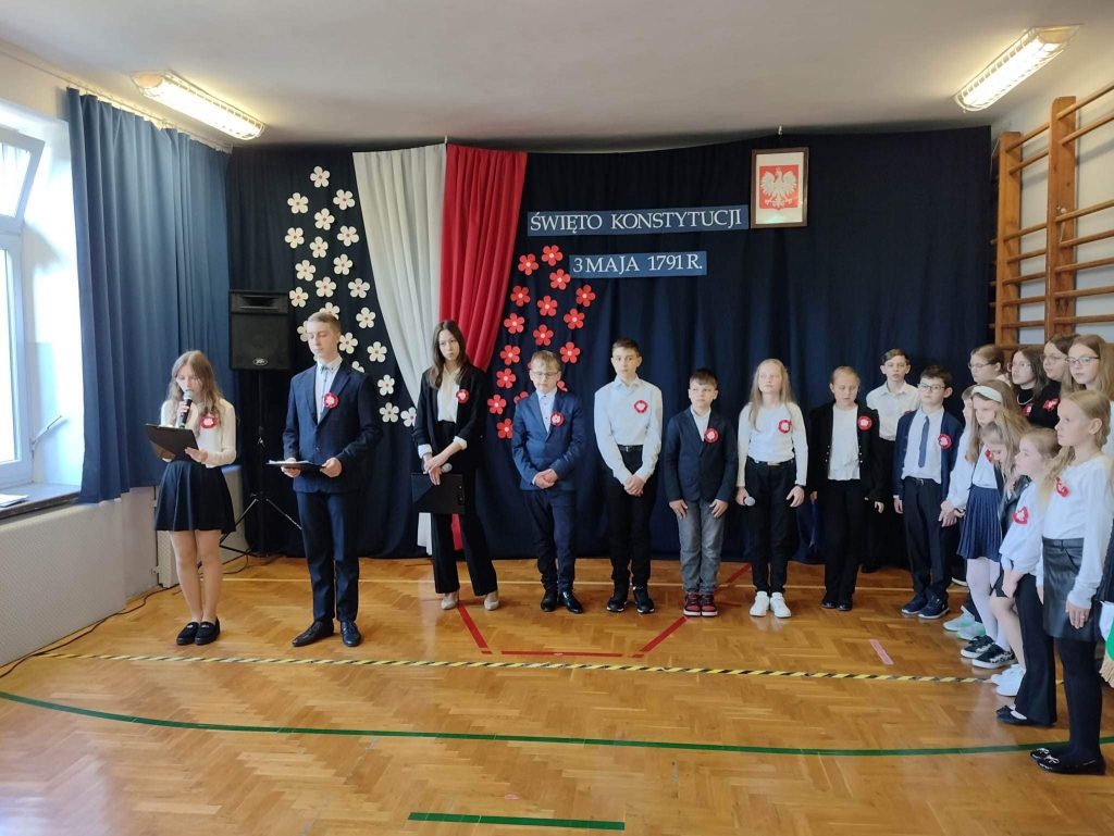 Uczniowie podczas uroczystości szkolnej w tle flaga Polski i napis ŚWIĘTO KONSTYTUCJI 3 MAJA 1791R.