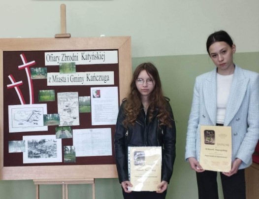 Uczennice z prezentują dyplomy po lewej stronie tablica z informacjami nt. Katynia