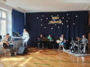 Uczniowie na sali gimnastycznej grają na instrumentach. w tle napis Dzień Talentów