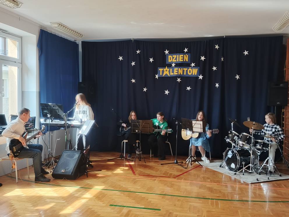 Uczniowie na sali gimnastycznej grają na instrumentach. w tle napis Dzień Talentów